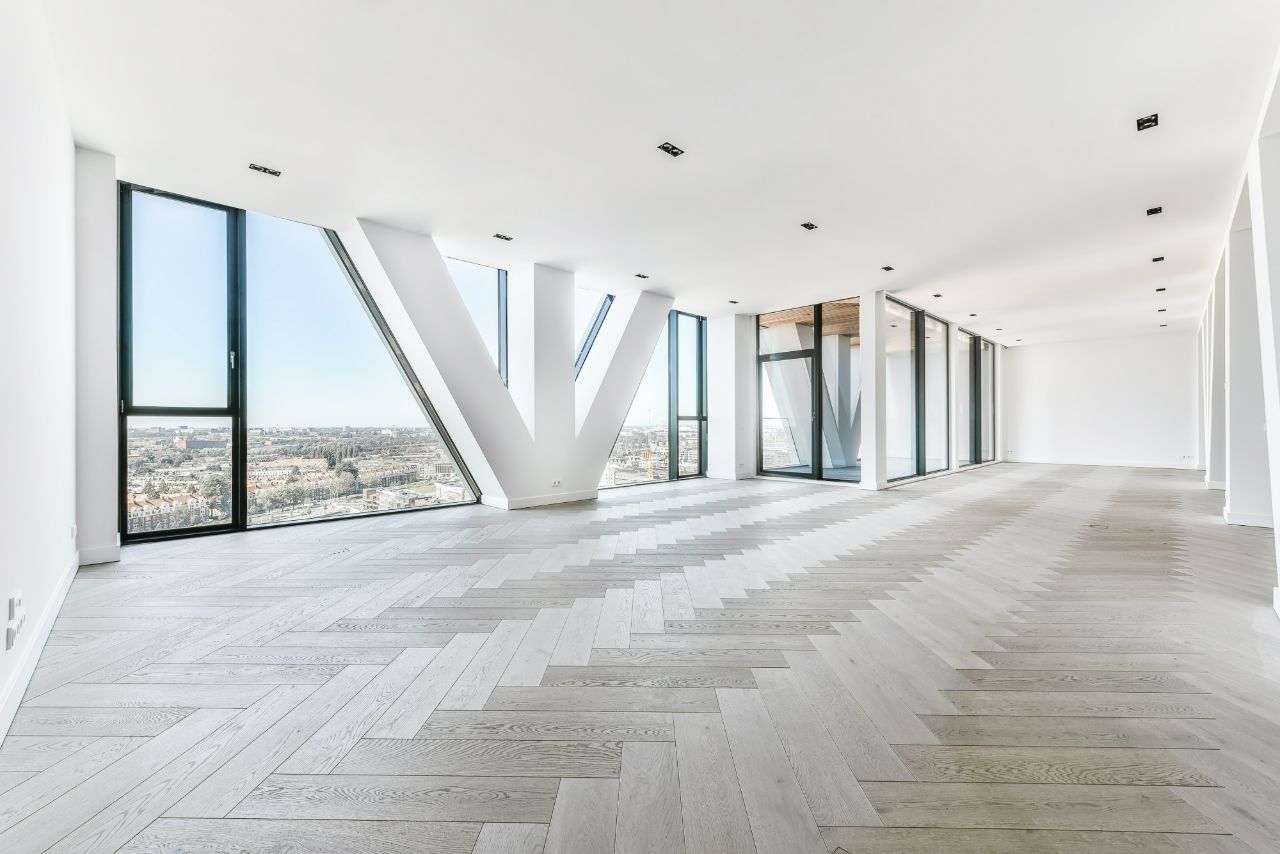 Bright empty penthouse with angled windows and pale herringbone wood flooring.