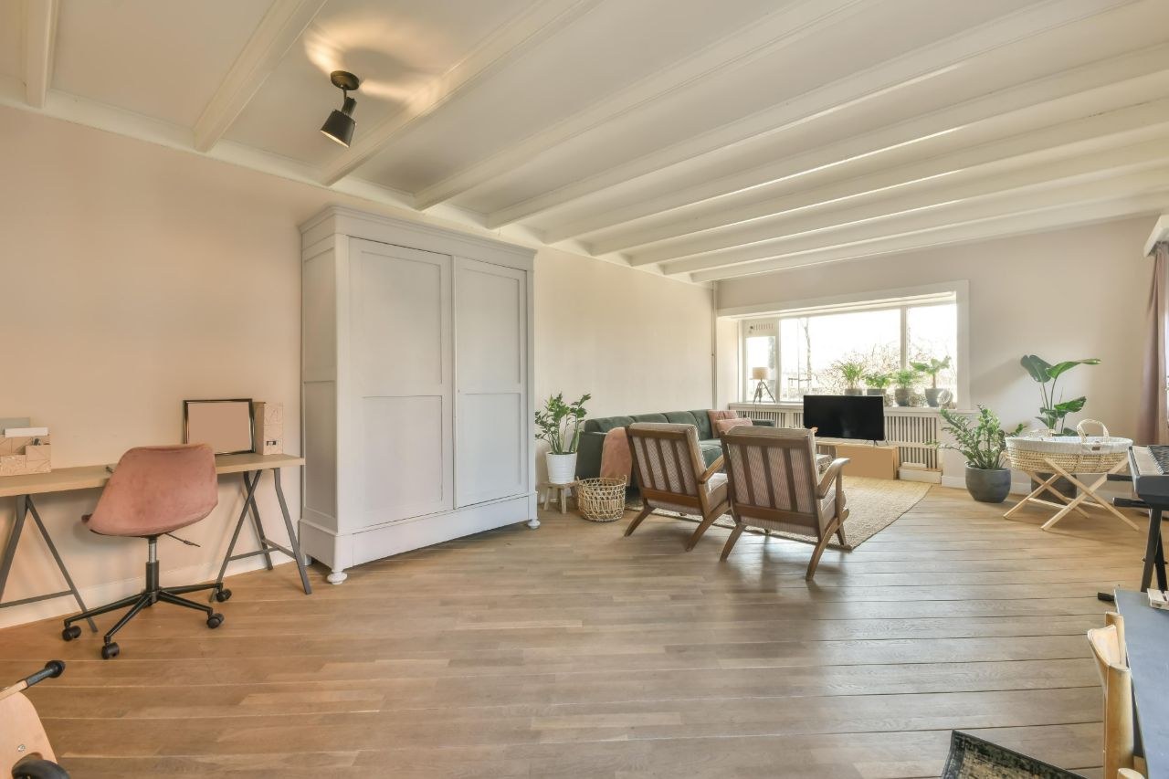 Lived-in living room with oak floorboards, a desk, two armchairs and plants by a wide window.