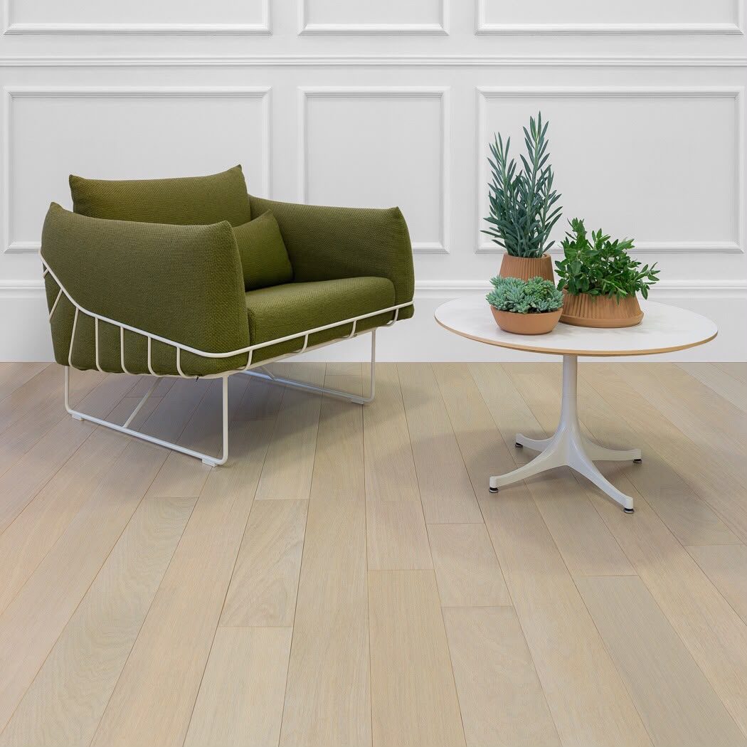Mistwood solid hardwood by Gold Flooring: pale, light-toned planks under a green upholstered armchair and a round white side table with potted succulents, against white panelled walls